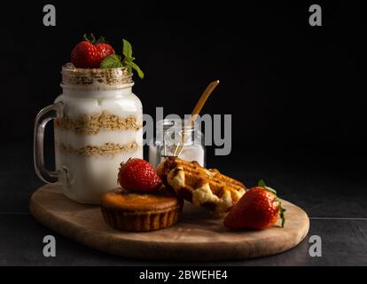 Homemade vegetarian breakfast, yogurt with strawberry and muesli with a waffle and apple cake on the side on a dark background. Stock Photo