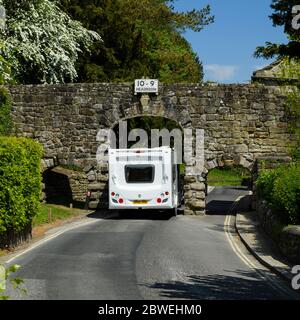 Caravan passing through narrow rustic stone archway spanning scenic country lane (tight squeeze) - B6160, Bolton Abbey village, Yorkshire, England, UK Stock Photo