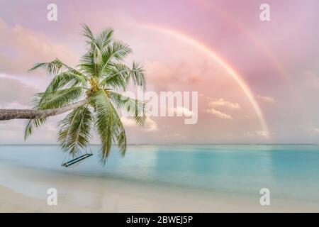 Perfect tropical landscape, sunset beach with palm tree and swing hanging under colorful rainbow. Romantic beach landscape Stock Photo