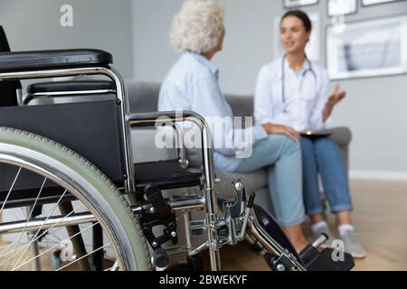 Physiotherapist elderly woman talking seated on sofa, focus on wheelchair Stock Photo