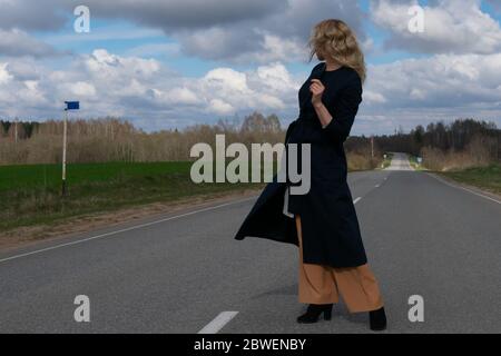 A middle-aged Caucasian woman in an elegant summer black coat stands alone near a highway in the countryside against a cloudy sky on a spring day. Cop Stock Photo