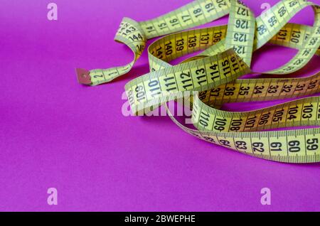 Yellow tailor measuring tape on a lilac background. Twisted measuring tape with decimal scale. Multitask background. Copy space. Selective focus. Stock Photo