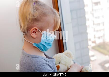 Caucasian little child girl wear protective mask looking outside through the window. Sad kid in quarantine room due to coronavirus. child staying at h Stock Photo