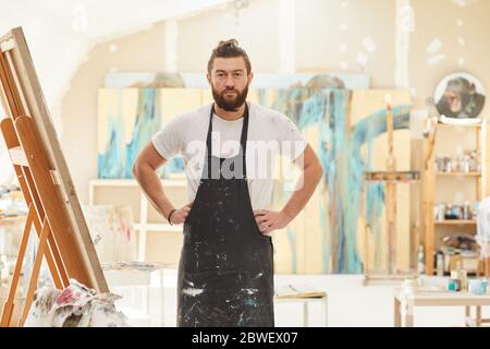 Portrait of mature bearded artist wearing apron and posing confidently looking at camera while standing in spacious art studio lit by sunlight, copy s Stock Photo