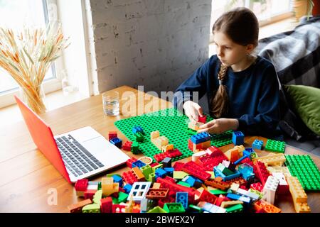 Girl playing with constructor at home, watching teacher's online tutorial on laptop. Digitalization, remote education concept. Technologies and devices. Man showing, giving online lesson. Artwork. Stock Photo