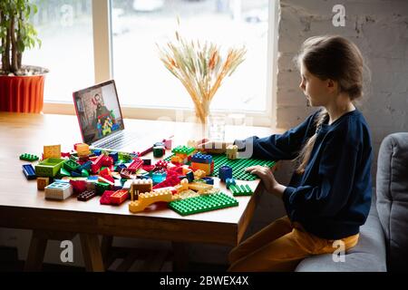 Girl playing with constructor at home, watching teacher's online tutorial on laptop. Digitalization, remote education concept. Technologies and devices. Man showing, giving online lesson. Artwork. Stock Photo