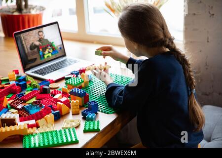 Girl playing with constructor at home, watching teacher's online tutorial on laptop. Digitalization, remote education concept. Technologies and devices. Man showing, giving online lesson. Artwork. Stock Photo