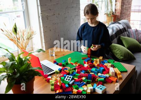 Girl playing with constructor at home, watching teacher's online tutorial on laptop. Digitalization, remote education concept. Technologies and devices. Man showing, giving online lesson. Artwork. Stock Photo