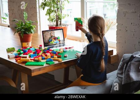 Girl playing with constructor at home, watching teacher's online tutorial on laptop. Digitalization, remote education concept. Technologies and devices. Man showing, giving online lesson. Artwork. Stock Photo