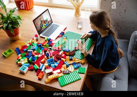 Girl playing with constructor at home, watching teacher's online tutorial on laptop. Digitalization, remote education concept. Technologies and devices. Man showing, giving online lesson. Artwork. Stock Photo