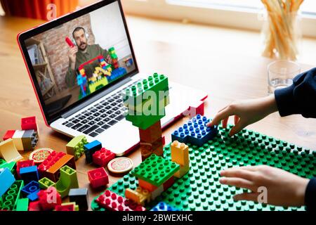 Girl playing with constructor at home, watching teacher's online tutorial on laptop. Digitalization, remote education concept. Technologies and devices. Man showing, giving online lesson. Artwork. Stock Photo