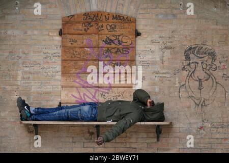 A boy lying on the wooden shelf of a newspaper kiosk closed in Italy, calls on his mobile phone, dressed in a hooded jacket that obscures his face. Stock Photo