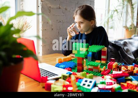 Girl playing with constructor at home, watching teacher's online tutorial on laptop. Digitalization, remote education concept. Technologies and devices. Man showing, giving online lesson. Artwork. Stock Photo