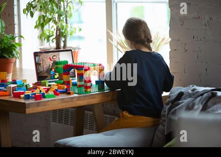 Girl playing with constructor at home, watching teacher's online tutorial on laptop. Digitalization, remote education concept. Technologies and devices. Man showing, giving online lesson. Artwork. Stock Photo