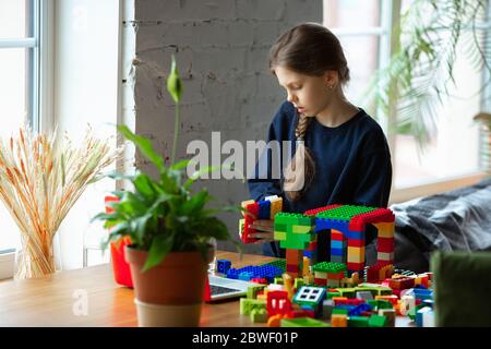 Girl playing with constructor at home, watching teacher's online tutorial on laptop. Digitalization, remote education concept. Technologies and devices. Man showing, giving online lesson. Artwork. Stock Photo