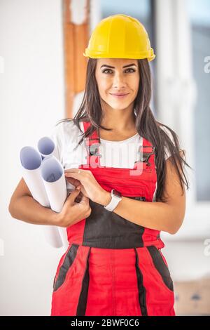 Good looking female construction planner holding blueprints dressed in working clothes and helmet inside the house. Stock Photo