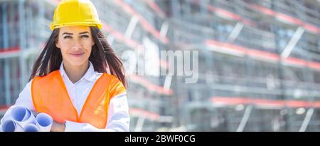 Female architect wearing orange safety vest and a yellow helmet holds rolled construction plans under her armpit in front of the unfinished building. Stock Photo