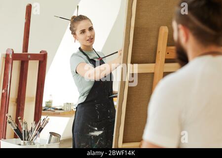 Portrait of young female artist looking at model while painting portrait in studio, copy space Stock Photo