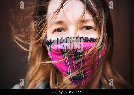 Closeup portrait of a young woman wearing a plaid handmade face mask with disheveled hair waving in the wind.