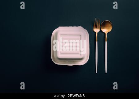 Healthy food concept: white burguer packaging closed with golden fork and spoon in cardboard lunch box to take to the office on black backgorund Stock Photo
