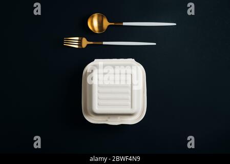Healthy food concept: white burguer packaging closed with golden fork and spoon in cardboard lunch box to take to the office on black backgorund Stock Photo