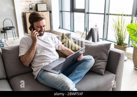 Young man using a laptop while talking on the phone sitting on the couch Stock Photo