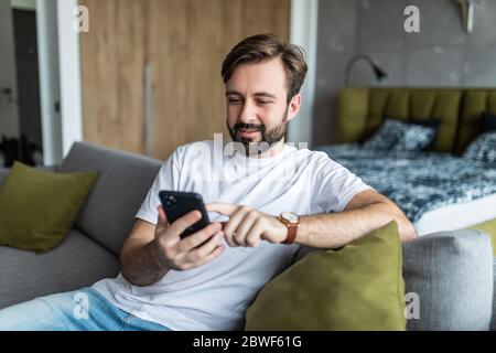 Millennial man sitting in living room on couch at home smiling chatting typing message using cellular. Stock Photo