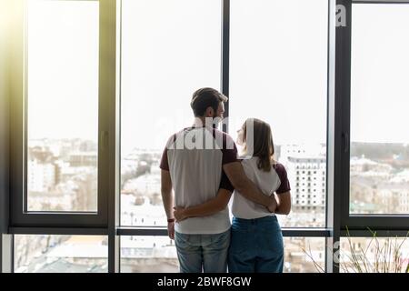 Young Beautiful Couple Stand Near Window Embrace, Happy Smile Hispanic Man And Woman Lovers Hug Back Rear View Stock Photo