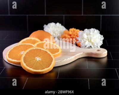 Round wooden cutting board with a handle with a fresh orange sliced up and arranged on it with 3 artificial white and orange flowers sitting behind it Stock Photo