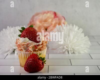 Strawberry shortcake cupcake garnished with a fresh strawberry on top and a strawberry in front of it with white and pink artificial flowers behind it Stock Photo