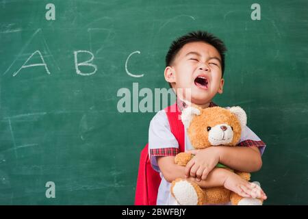 Back to School. Happy Asian funny cute little child boy from kindergarten in student uniform with school bag smiling and hugging teddy bear on green s Stock Photo