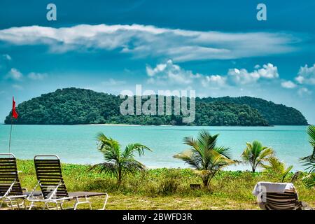 Small palm trees and deck chairs looking at a tropical island in the azure sea under the blue sky on the shores of the sandy beautiful exotic and stun Stock Photo