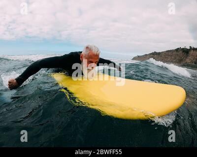 Senior man doing surf with longboard riding a wave - Mature person having fun doing extreme sport - Joyful elderly concept - Focus on his face Stock Photo