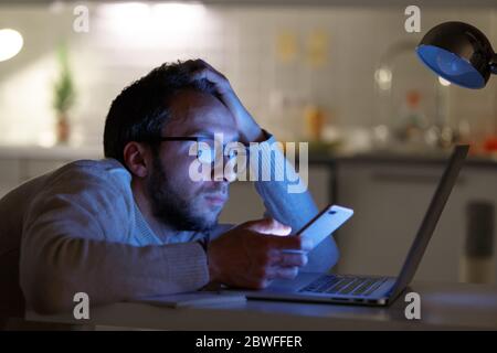 Sleepy tired man designer in eyeglasses using smart phone, working/typing on laptop computer at night home. Freelance, overwork, deadline, remote work Stock Photo