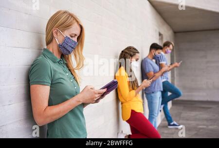 Young people wearing face safety masks using smart mobile phones while keeping social distance during coronavirus time - Technology and covid-19 sprea Stock Photo