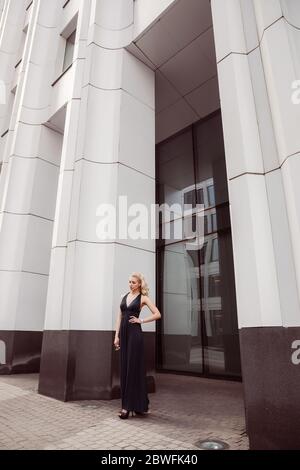 Pretty blonde female in the city is standing in a portal of a large white building she is wearing sunglasses and a long black shoulderless dress Stock Photo