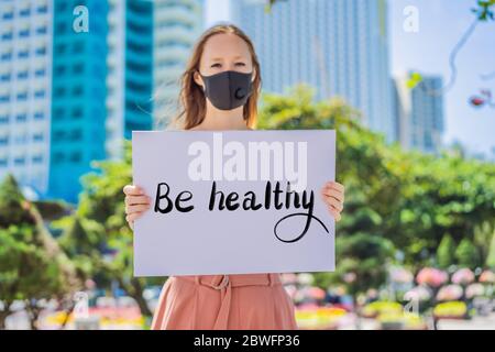 woman in medical mask prevents coronavirus disease holds a poster BE HALTHY Hand written text - lettering isolated on white. Coronovirus COVID 19 Stock Photo