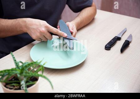 Close-up strong male hands sharpen a kitchen metal knife with a grindstone. Home household knife sharpening. Stock Photo