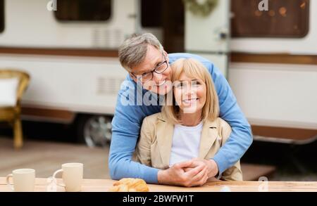 Smiling mature man hugging his beautiful wife in front of camping vehicle outdoors Stock Photo