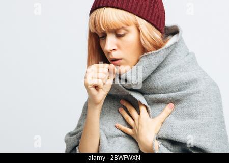 Cute sick young teen woman in red hat, wrapped in warm scarf coughing, closed eyes.Female feeling the first symptoms of illness.Bronchitis, upper resp Stock Photo