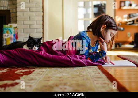 a child curls up on floor in a sleeping bag with cat doing schoolwork Stock Photo