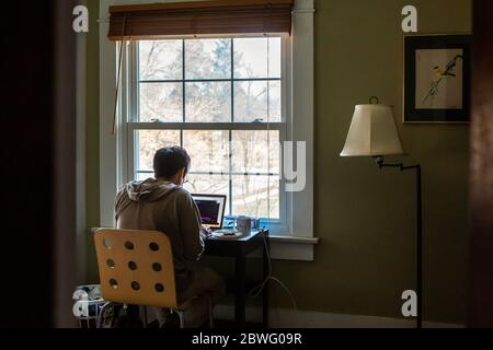 A man sits at desk in front of window in bedroom working on a computer Stock Photo