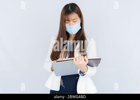 Asian business woman wearing a medical face mask and looking at the notebook that she holding over grey background. Back to the normal concept. Stock Photo