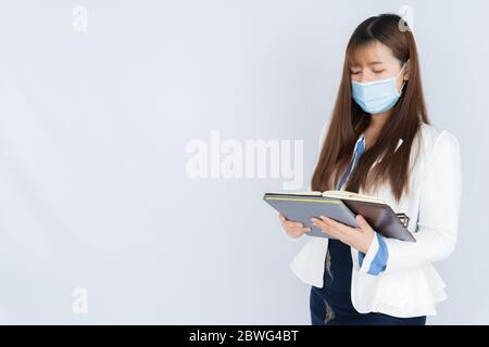 Asian business woman wearing a medical face mask and looking at the notebook that she holding over grey background. Back to the normal concept. Stock Photo