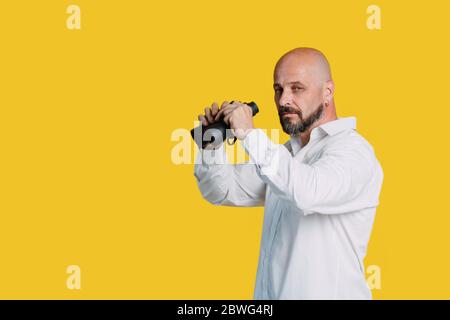 Handsome serious middle-aged man in a white shirt smiles and holds black binoculars on a yellow background with copy space search concept  Portrait of Stock Photo