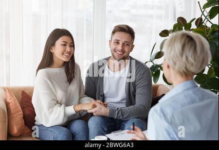 Smiling Multiethnic Spouses Holding Hands Reconciling Sitting In Psychotherapist's Office Stock Photo