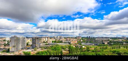 Large low clouds fill the sky over a sunny city with tall apartment buildings and green parks in a panorama with cityscape and horizon.
