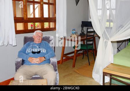 senior man asleep in chair, nap, resting, guest room, mosquito netting, bed, 2 windows, Ngorongoro Farmhouse Valley Lodge; Tanzania, Africa; MR Stock Photo