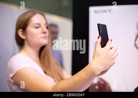 019: a a young blondie lady holds Sony Xperia 5 Phone black and looks into it doing selfie. front view, soft focus Stock Photo