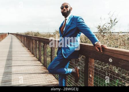A handsome brutal bald bearded African man in sunglasses and a blue fashionable costume with a tie is leaning against a wooden pedestrian bridge Stock Photo